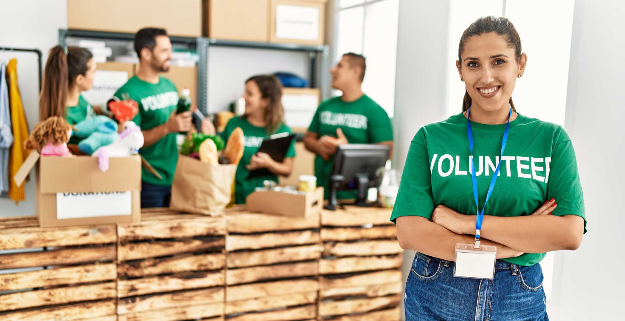 A smiling volunteer in a 'Volunteer' t-shirt stands with arms crossed in a donation center, with other volunteers sorting donations behind her.